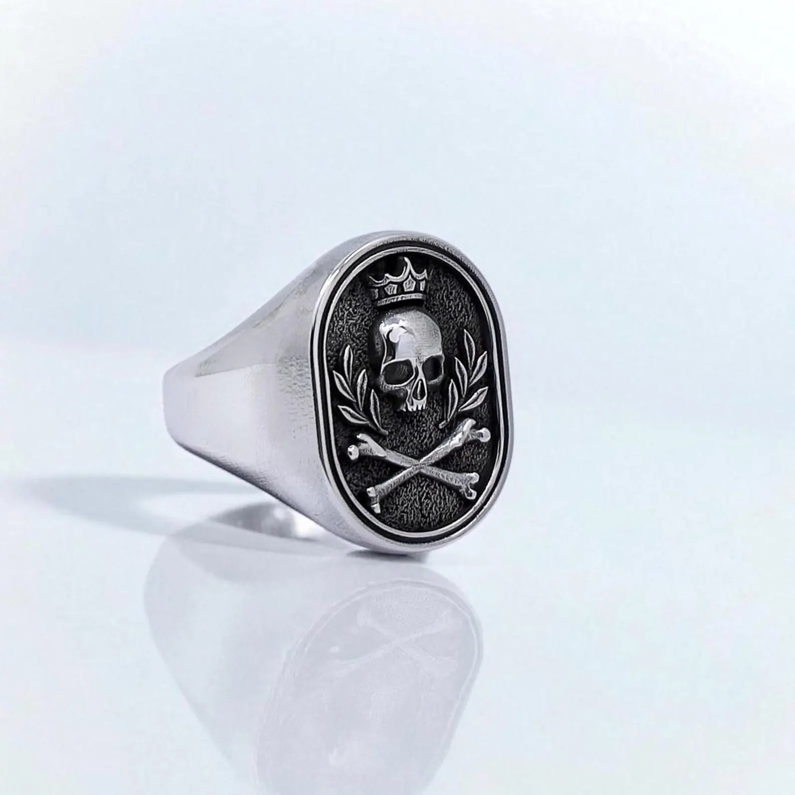Front-facing view of a sterling silver signet ring featuring a crowned skull and crossbones design, surrounded by laurel branches. This handcrafted gothic jewelry piece exudes rebellious energy and bold personality, perfect for men who embrace punk, rock, or biker aesthetics.