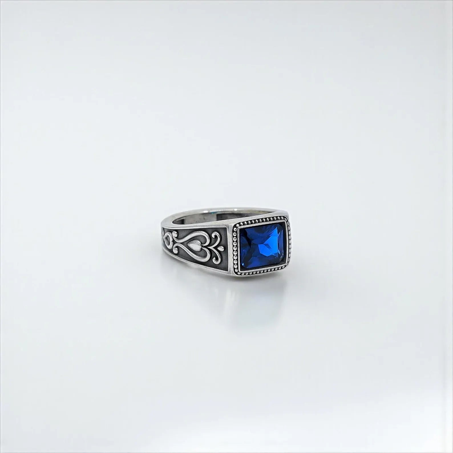 Stylized shot of a men's sterling silver ring showcasing a vivid sapphire blue emerald-cut zircon stone, surrounded by vintage gothic patterns – a refined and powerful accessory for everyday or special occasions.
