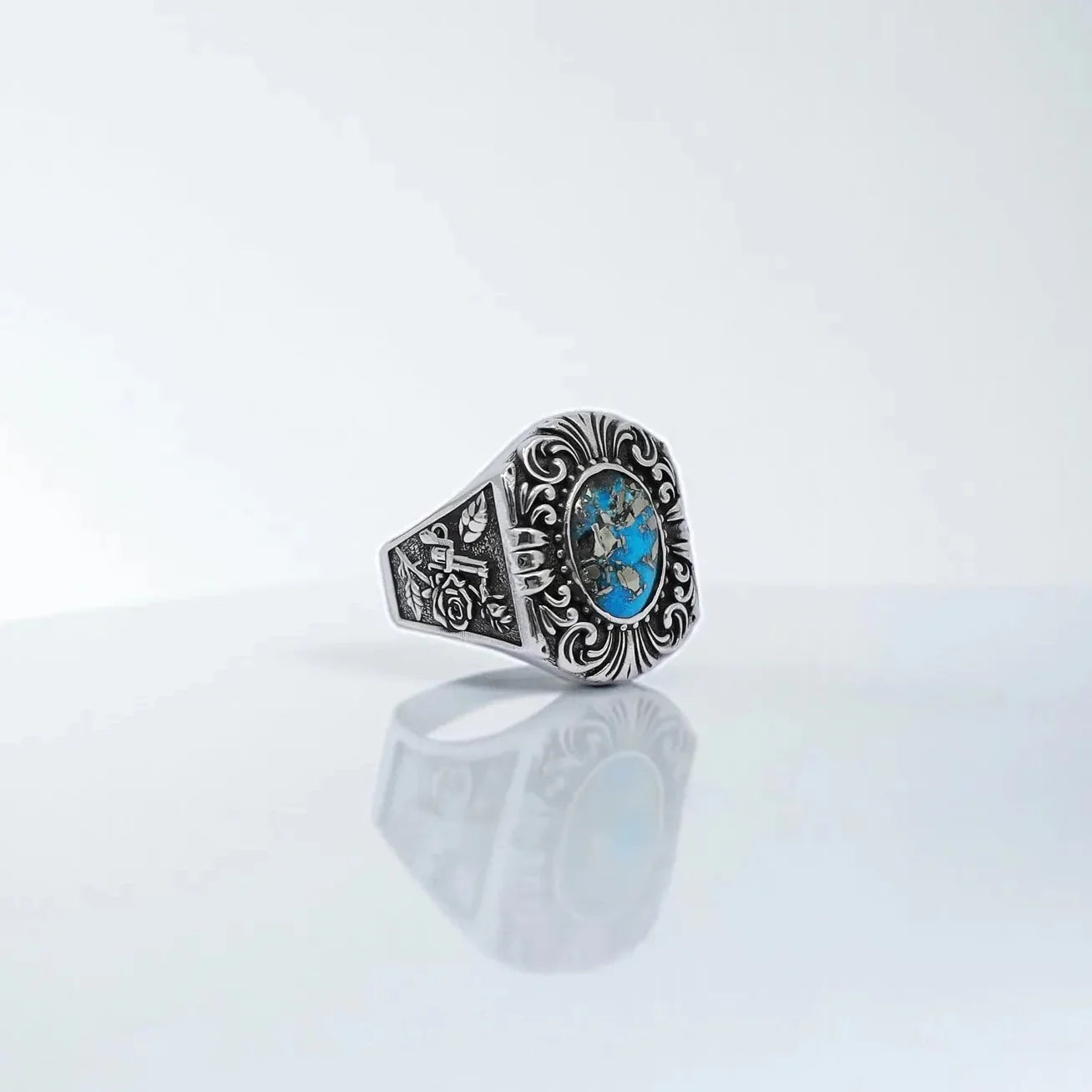 Close-up image of a handcrafted sterling silver men's ring featuring an oval turquoise gemstone, surrounded by ornate gothic scrollwork and engraved rose and gun motifs – a bold western-style statement piece.