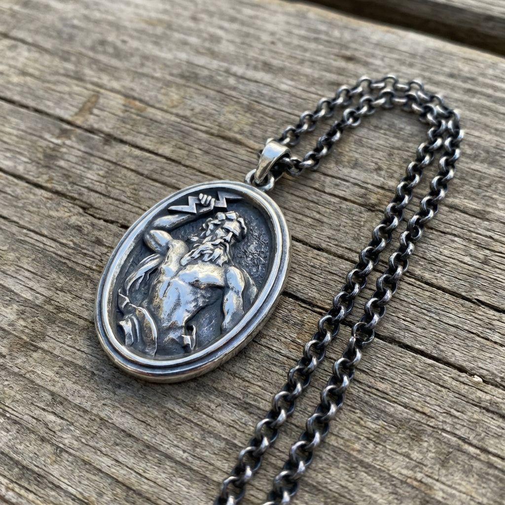 May include: A silver-toned necklace featuring an oval pendant with a detailed relief of a mythological figure. The figure is depicted with a beard and holding a lightning bolt. The chain is a matching silver color.
