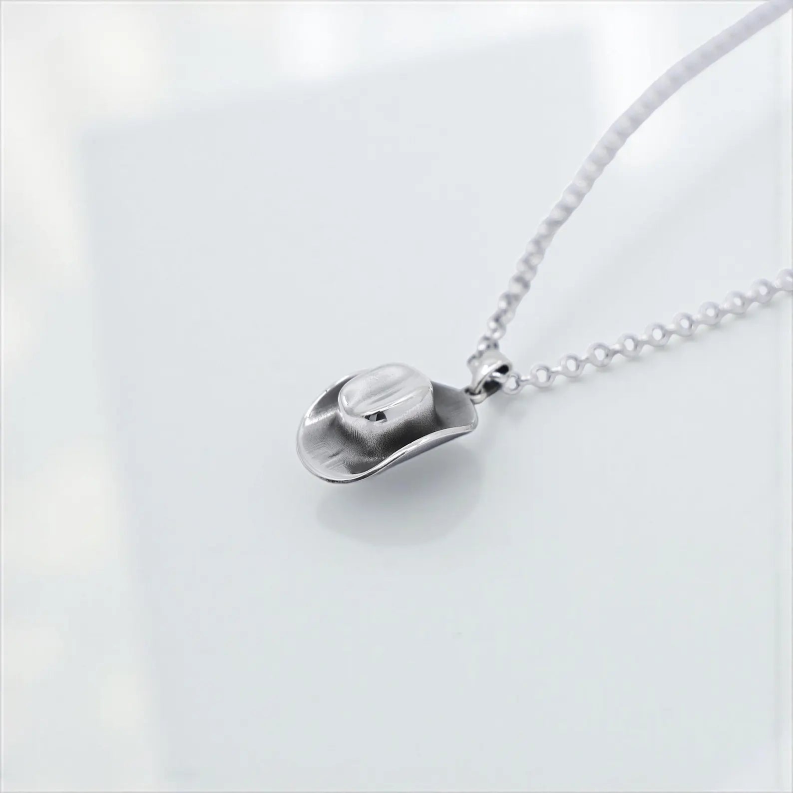 Close-up of a handcrafted sterling silver cowboy hat pendant necklace, featuring a detailed western hat charm with brushed texture, suspended on a thick silver chain and photographed against a clean white background.
