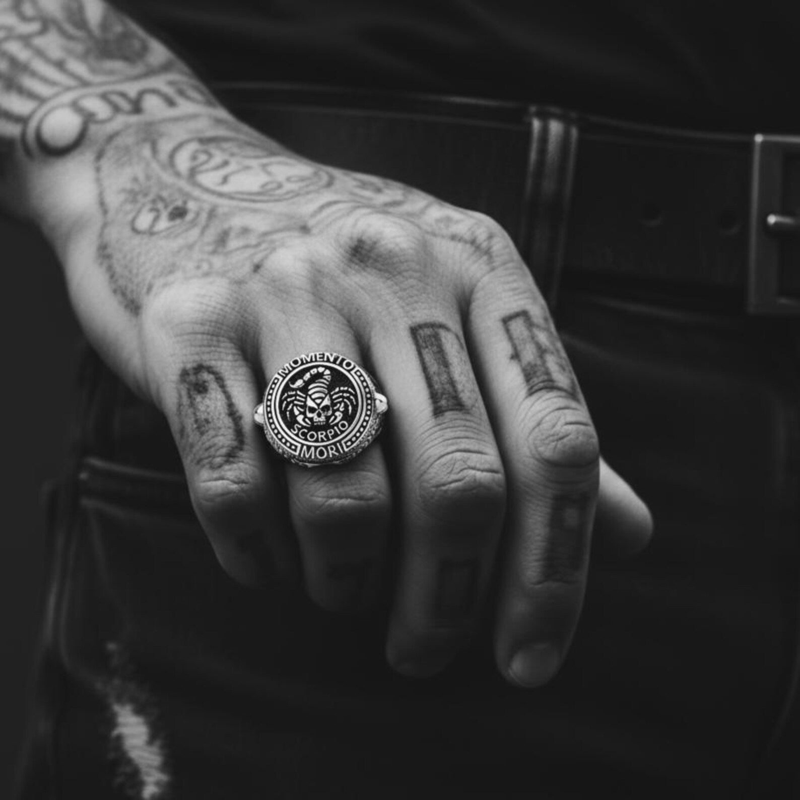 May include: A silver ring with a detailed design featuring a scorpion, skull, and the words "MOMENTO MORI SCORPIO". The ring is worn on a hand with tattoos.